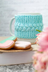 A white cup of tea in a knitted blue case stands on a book and pink flowers, tulips and gingerbread on a texture table. Spring concept