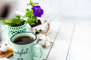 Close-up of a cup of tea on a wooden white table with blurred background, front blur. Still life with flower, book, teaspoon, cotton box. Spring breakfast. Copy space.