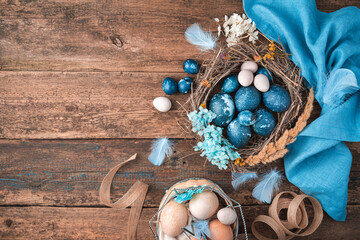 Obraz na płótnie Canvas Marble-blue Easter eggs in a wicker nest with feathers and flowers next to a blue napkin and a basket of pastel eggs. Top view, horizontal.