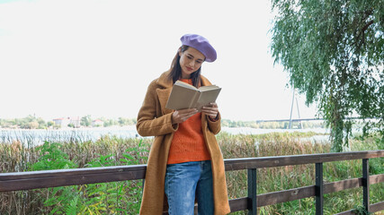 young woman in trendy autumn clothes reading novel on bridge in park