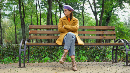 trendy displeased woman looking away while sitting with crossed arms on bench in park