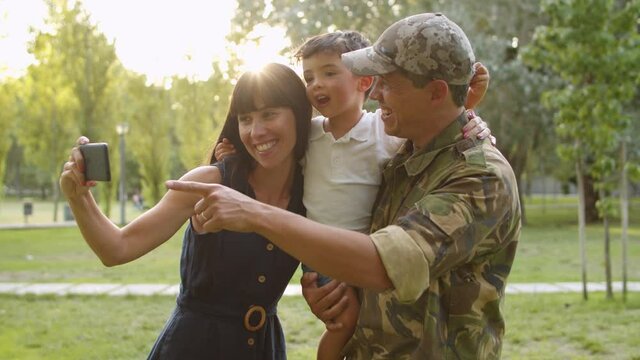Happy military family couple with kids celebrating dads returning, enjoying leisure time in park, taking selfie on smartphone. Family reunion or returning home concept