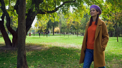 happy woman in stylish autumn outfit strolling in park with hand in pocket