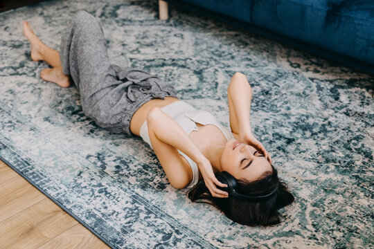 Young woman listening to music in wireless headphones, lying on the floor on carpet, smiling.