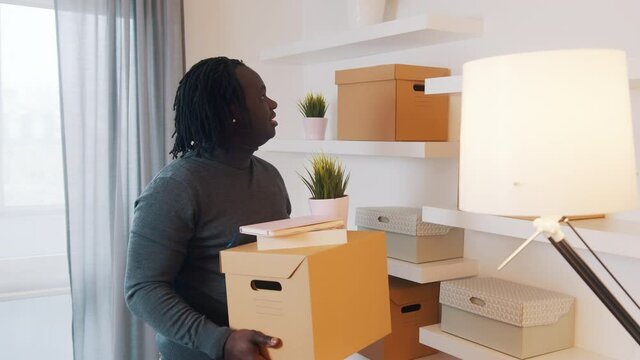 Young african american man moving in to the new appartment. Holding carboard boxes and plant. High quality 4k footage