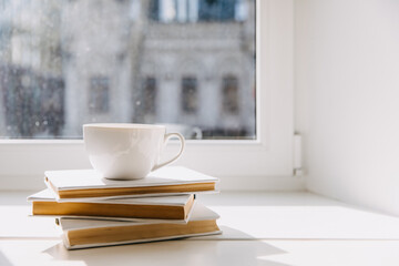Blank white cup of coffee or tea on stack of books by the window.