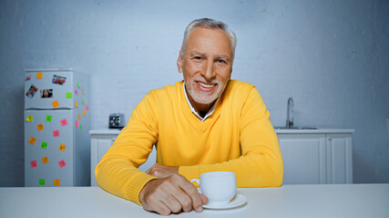 Cheerful senior man looking at camera near coffee cup in kitchen