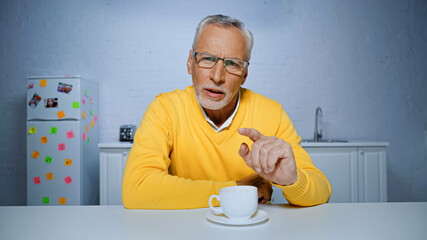 Elderly man in eyeglasses pointing with finger at camera near cup of coffee in kitchen