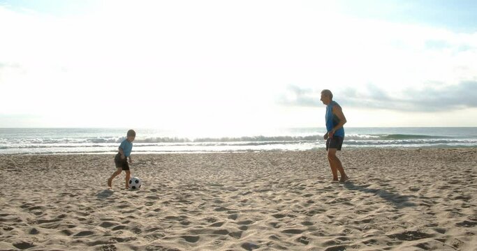 Football soccer game on the beach. Man playing football with his little boy on the sandy sea beach, 4k slow motion