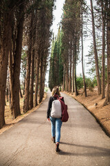 A woman hiker with backpack walks down an alley of cypresses view from behind on a warm spring day.
