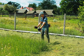 A man mows the grass with a trimmer on a sunny day. Photo of a farmer mowing grass in a village in the backyard. A quiet scene. Front view of a man with a petrol mower.    
