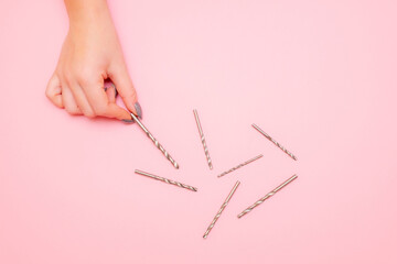 a woman's hand picks up a drill bit. a pile of drills lies on a pink background