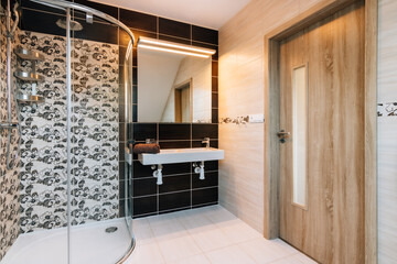 Beautiful small bathroom in attic of family house.Bathroom has large glass shower,double basin with large illuminated mirror and wooden entrance door.Tiling differs a lot in terms of color or texture.