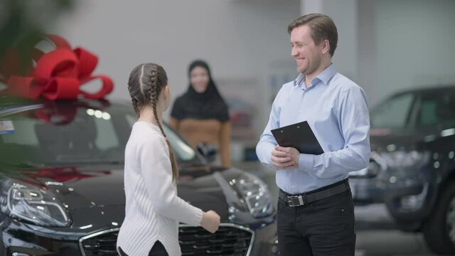 Cheerful car dealer and joyful little girl giving high five in slow motion in car dealership. Happy employee and client rejoicing new vehicle buying in showroom. Blurred woman in hijab at background.