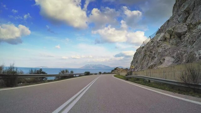 Coastal rocky scenery POV vehicle drive, mediterranean coast curvy asphalt road with concrete  land slide barrier, blue sky cloudy horizon, point of view car travel