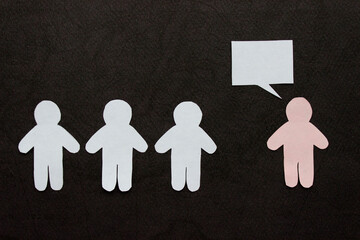 Silhouettes of people cut from white and pink paper on a black background. A group of people stands in front of one person (pink figure with speech-bubble). Communication, leadership, teamwork concept