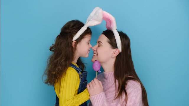 Happy family play together, keep noses together on purple Easter egg, enjoy pleasant moments of preparation to spring Christian holiday. Lovely daughter spends time with mother, isolated on blue wall
