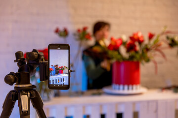 Florist workplace on the background of a white brick wall. A florist creates a flower arrangement in a red box and records an educational video on a smartphone. Online lesson, video conference
