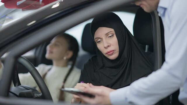 Confident gorgeous woman in hijab signing purchase and sale agreement sitting on driver's seat in showroom. Portrait of wealthy beautiful lady buying automobile in dealership with daughter.