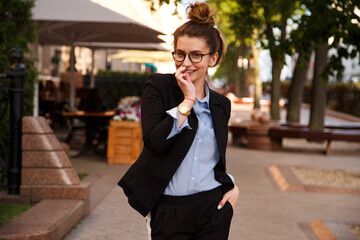 Street portrait of charismatic young woman in formal suit and eyeglasses walking on street. Outdoor photo of smiling shy office girl with brown hair tied in a bun