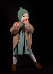 Portrait of little girl (toddler), dressed in cozy vintage clothes. She poses and nibbles an apple. Baby teeth, vitamins for babies. Studio isolated at black background.