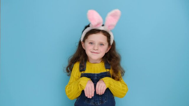 Funny preschooler girl keeps hands as if rabbit, wears soft bunny ears, has friendly look, smiles cheerfully, ready to congratulate children with coming Easter, models over blue wall. I am lovely hare