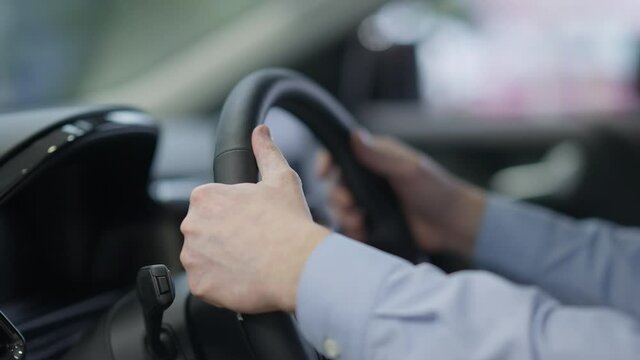 Close-up of male Caucasian hands on car steering wheel. Unrecognizable successful businessman choosing vehicle in dealership. Rich confident man buying automobile in showroom.