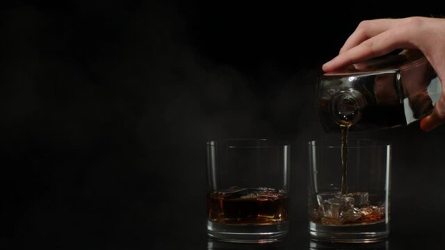 Hand of barman pouring golden whiskey, cognac or brandy from bottle into glasses with ice cubes on black background. Bartender pouring glass of alcohol drink bourbon, rum. 4k, slow motion, close-up