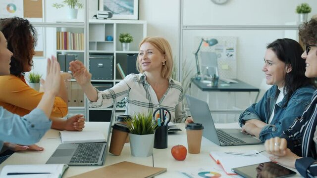 Joyful ladies multi-ethnic team talking and doing high-five enjoying professional achievement together sitting at desk indoors in workplace. Business and feminism concept.