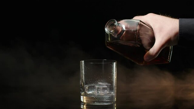 Barman pour golden whiskey, cognac, brandy from bottle into drinking glass with ice cubes on black dark background. Bartender pouring tasty alcohol cocktail drink bourbon, rum in slow motion. Close-up