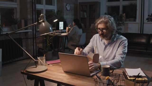 Medium shot of adult man working on laptop late sitting in dark office and eating chinese food from carton box