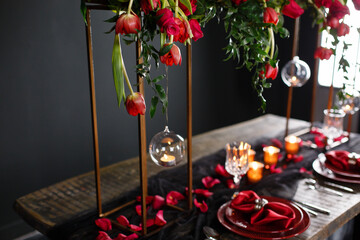 Table setting with red accents. Red plate on wooden table. Beautifully and festively served table with candles and fresh red rose flowers. Romantic date. Plate with red napkin and brooch - Powered by Adobe
