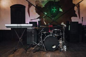 On stage there is a dark drum kit with cymbals and a microphone stand, two trumpets (alto, bass), a tambourine and a keyboard synthesizer. Spears, shield and sword on the wall.Jazz band live concert.
