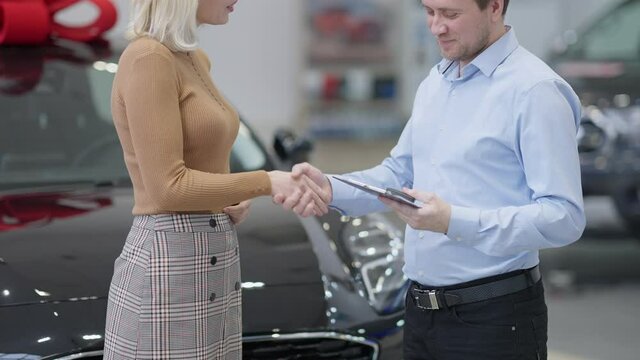 Unrecognizable dealer shaking hands with buyer and passing car key in showroom. Side view of happy slim Caucasian woman buying vehicle in car dealership. Luxury and lifestyle.
