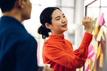 Casual creative happy two smile asian business people planning strategy analysis and brainstorm putting post it stickers note to share idea for startup project on glass board at modern office