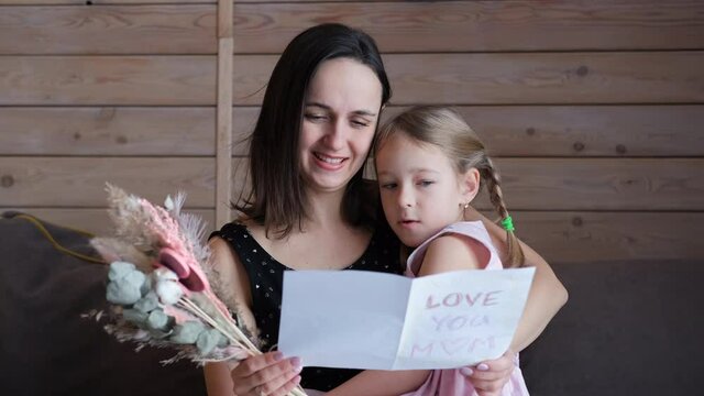 Young Mother Reading Greeting Card from her Little Daughter on Mothers Day. Slow Motion. Family, Togetherness and Happy People Concept