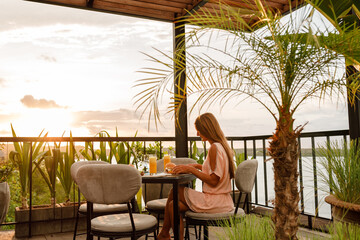 Attractive young woman sitting in outdoor restaurant by the sea and eat burger during sunset