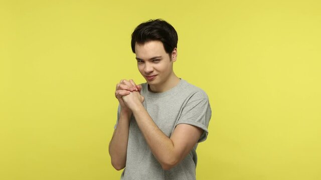 Young cunning guy in casual gray t-shirt drumming fingers, thinking devious evil idea, genius plan to prank friend, smirking and conspiring. Indoor studio shot isolated on yellow background