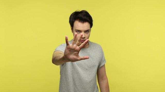 Young strict brunette man in casual gray t-shirt raising hand showing stop gesture, warning of troubles, way prohibited dangerous to go. Indoor studio shot isolated on yellow background