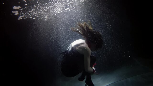 A dark-haired woman in red handcuffs is swimming deep under the water on a dark background, she is curled up with her butt up and creates a lot of bubbles.
