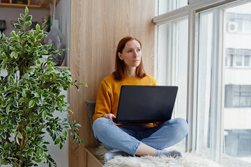 An employee or student in a yellow sweater is sitting by the window with a laptop. It is cozy and convenient to work remotely.