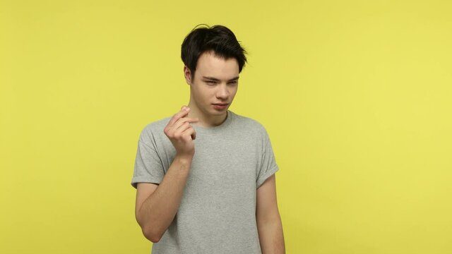 I need cash. Sly young man in casual style gray t-shirt showing money gesture and smiling, pretending to scattering money, planning to become rich. Indoor studio shot isolated on yellow background