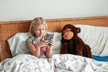 Cute girl sitting in bed in morning and texting talking at social media on smartphone cellphone. Child kid typing messaging on a mobile phone. Chat talk with friends online on Internet. Video chat.