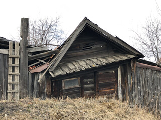 An old rickety wooden hut with boarded-up windows. There is dry grass and trees all around. The house is falling apart from old age, the boards are darkened.