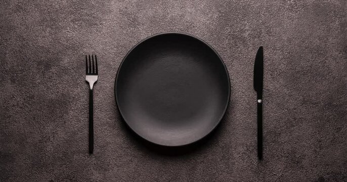 black dishes on a dark background with a rotation effect, top view. A plate and a spoon and fork on the black table