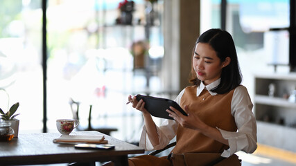 Elegant young female using digital table while sitting in modern cafe working on business project.