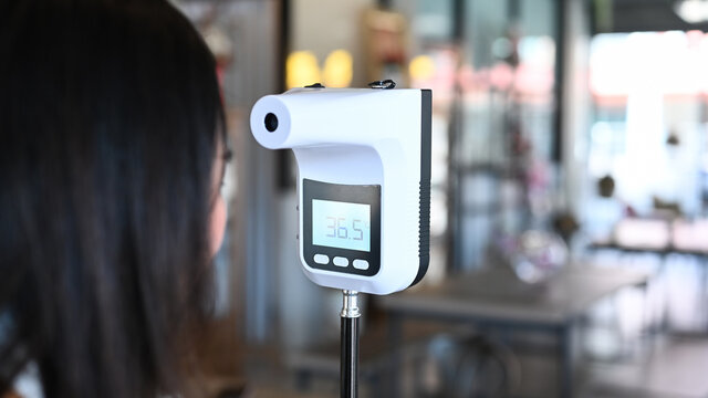 Young Asian woman body temperature checking before access to building by infrared forehead thermometer.
