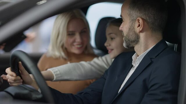 Confident rich man sitting on driver's seat talking with wife and daughter. Side view portrait of confident Caucasian wealthy father and husband choosing vehicle in dealership with family.