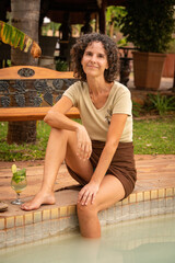 Naklejka premium Smiling brunette sitting with cocktail by pool