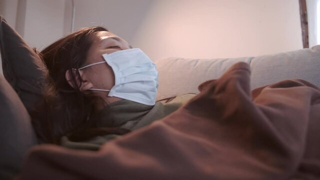 Young Asian woman suffering from coronavirus has black eyes chills all over her body gets cold and uses a mask and blanket to keep warm on the sofa in her separate living room and quarantine
